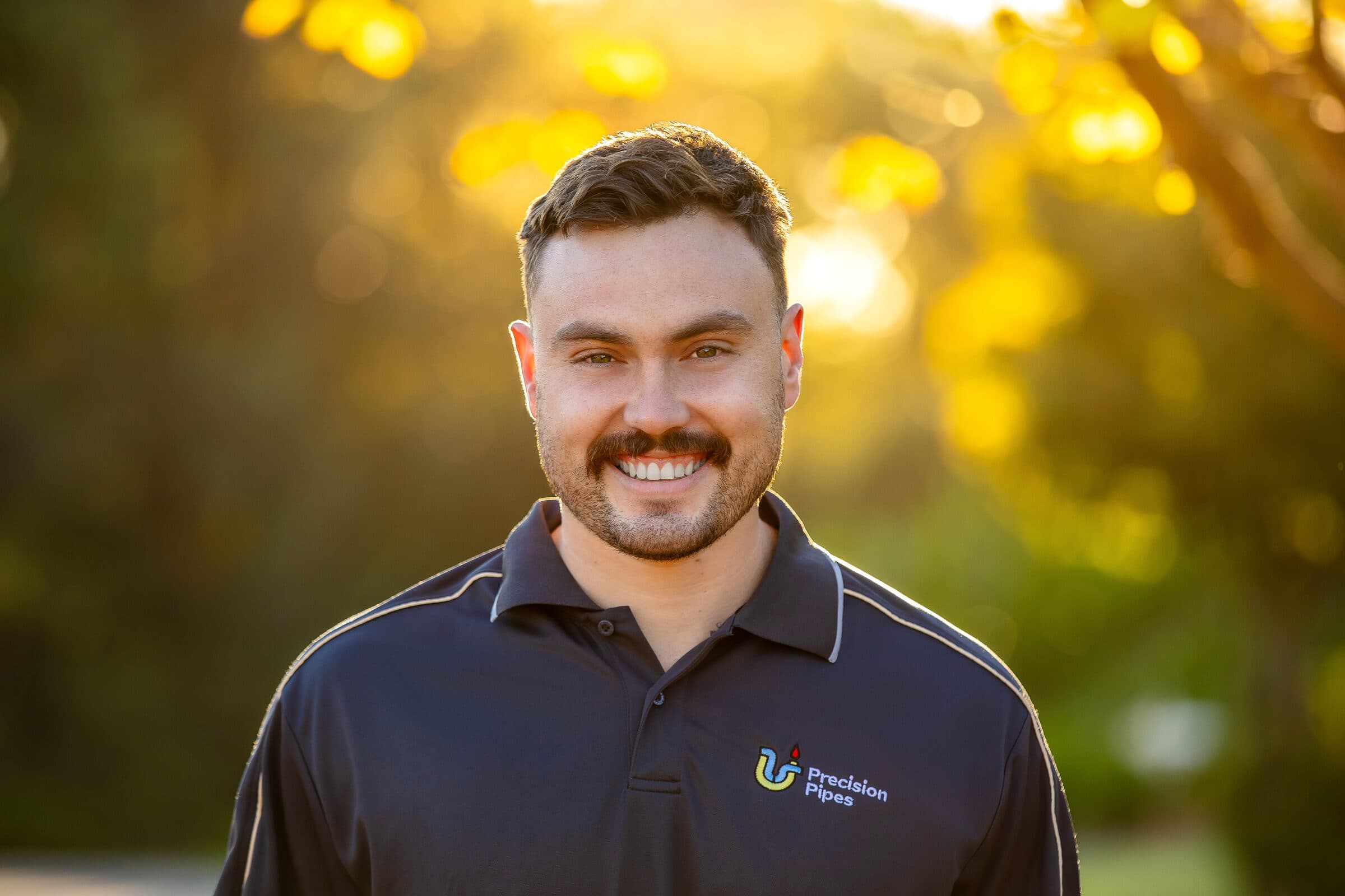 Rohan, the owner of Precision Pipes, in a branded navy polo, photographed on a tree-lined street at golden hour